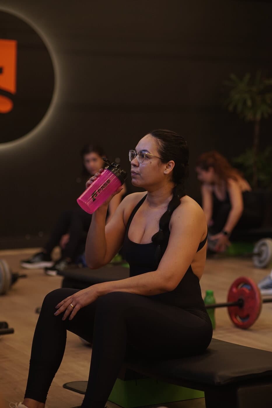 Aluna sentada no box bebendo água, com a turma treinando ao fundo.