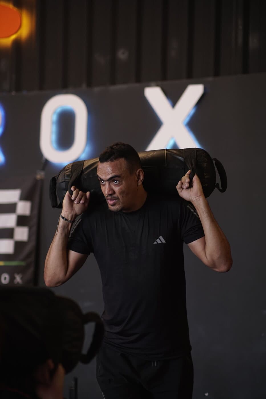 Aluno carregando o sandbag nos ombros, letreiro Hyrox ao fundo.
