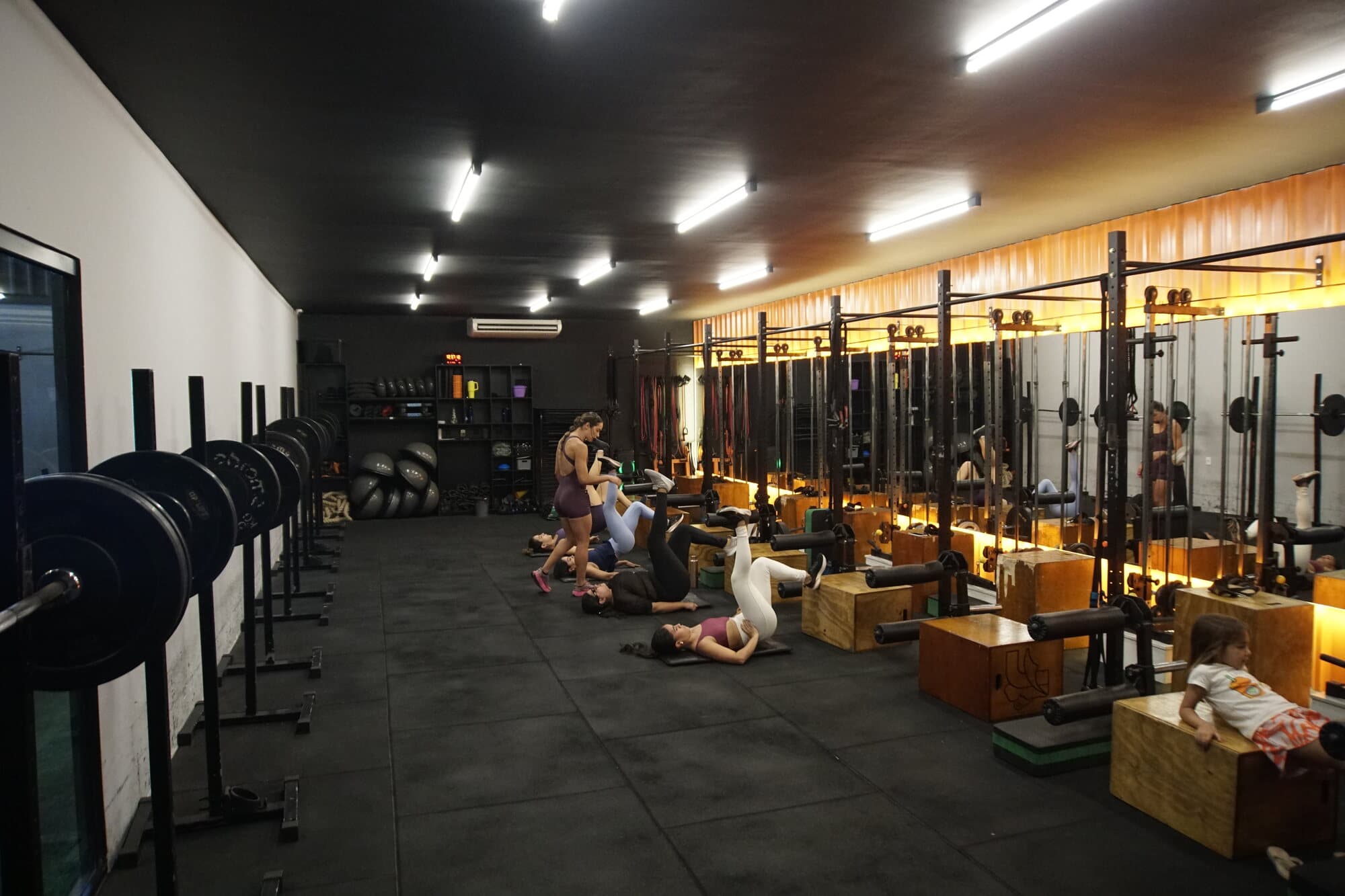 Sala de força da FS Studio: fileira de racks com barras carregadas, caixas de salto e alunos treinando no chão.