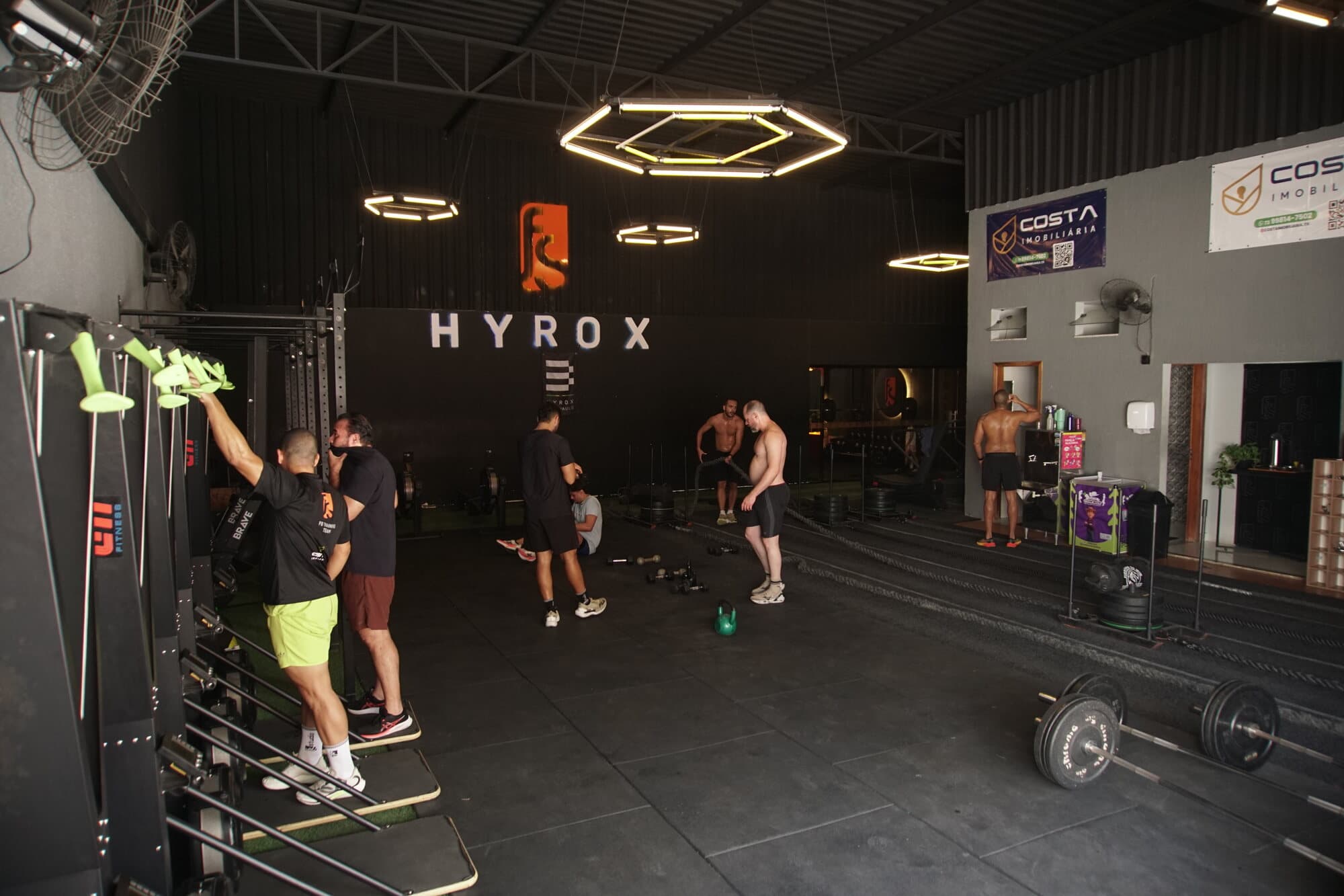 Salão principal da FS Studio durante uma aula: luminárias hexagonais, letreiro Hyrox na parede, alunos nos ski ergs e no piso.