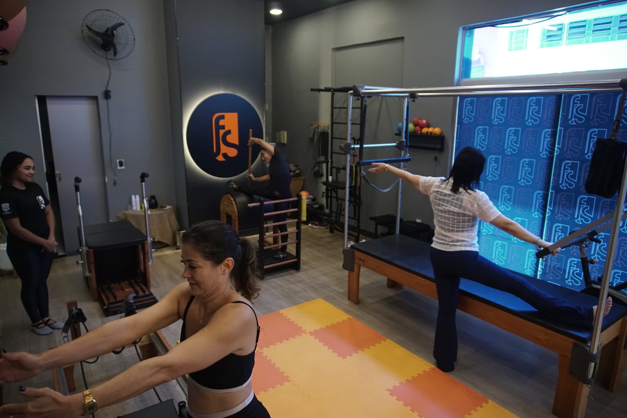 Sala de Pilates da FS Studio durante a aula: aluna no reformer, professora ao lado e o logo circular iluminado na parede.