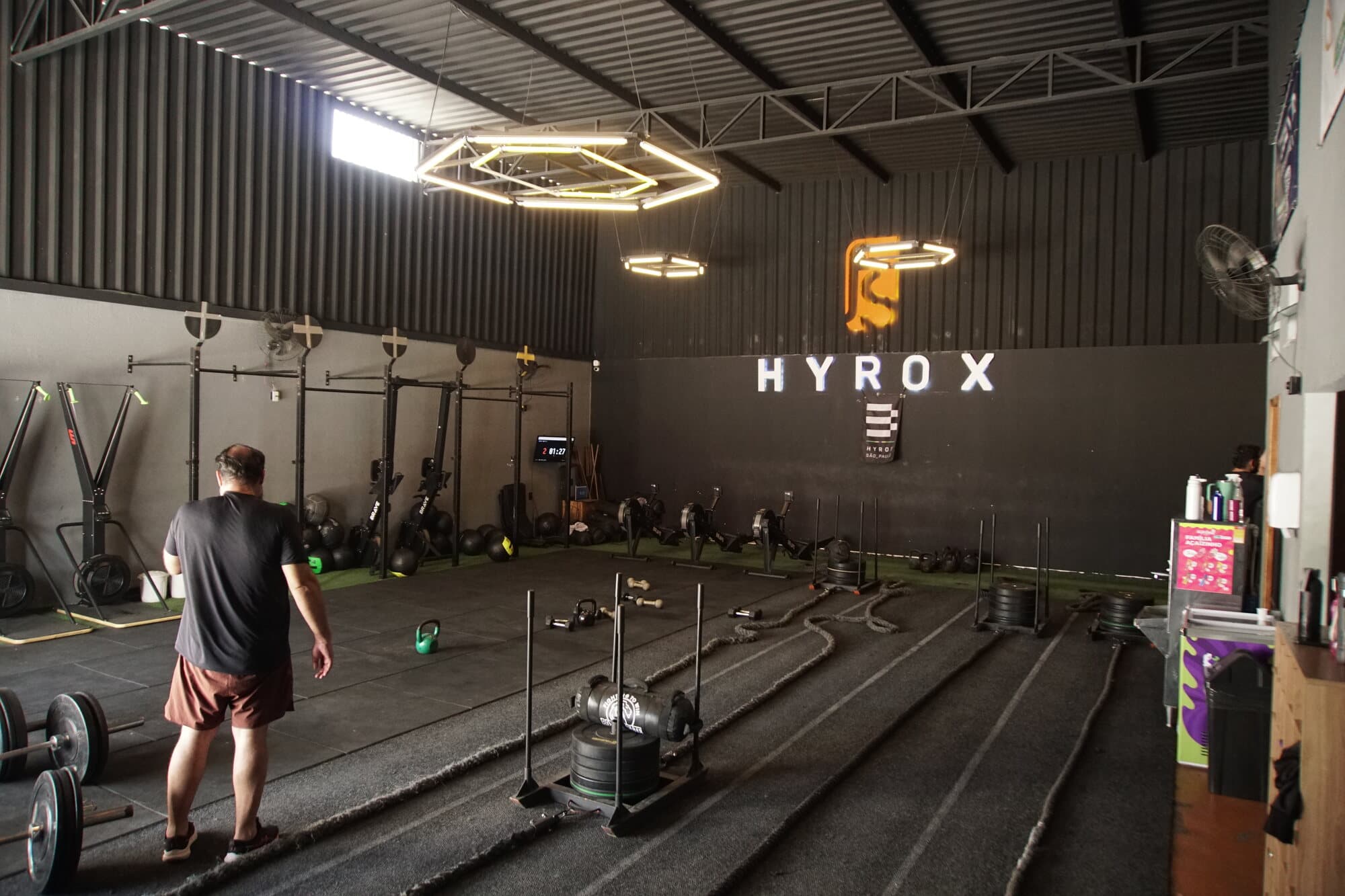 Galpão da FS Studio com o trenó carregado na pista de grama sintética, letreiro Hyrox ao fundo.