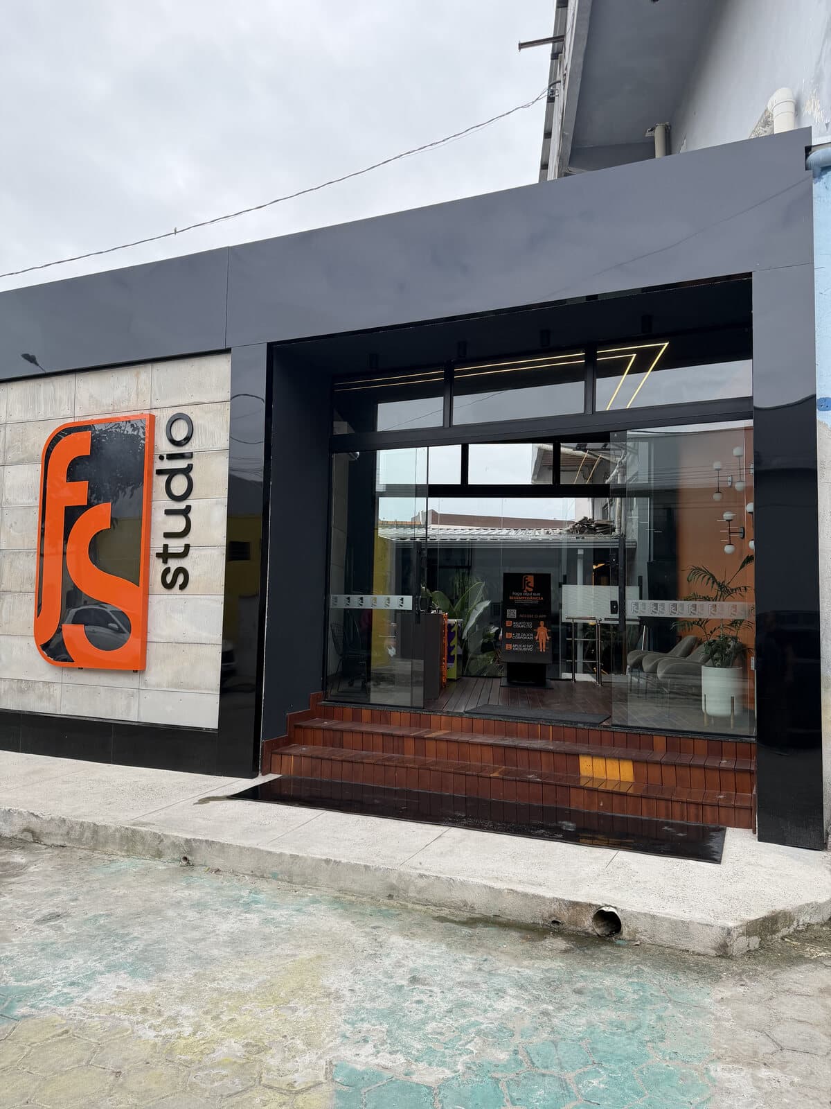 Fachada da FS Studio: logo laranja sobre a parede de pedra, letreiro studio e entrada de vidro com escada de tijolo.